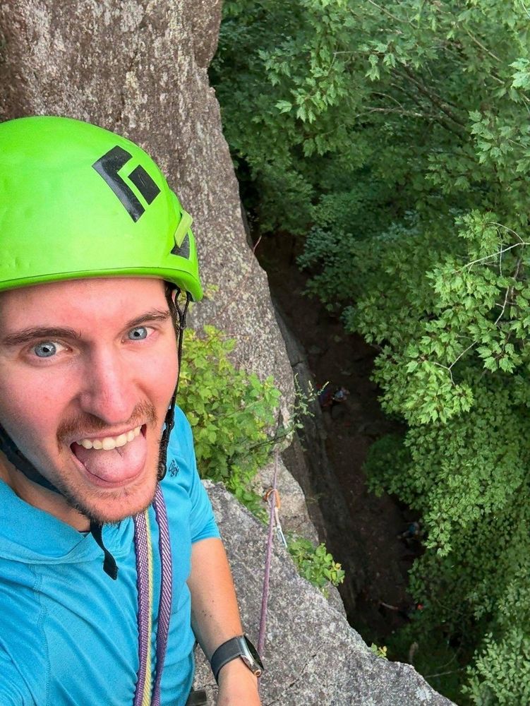 A climber wearing a bright green helmet and blue shirt smiles while standing on a rock face surrounded by lush green trees and secured with climbing ropes.