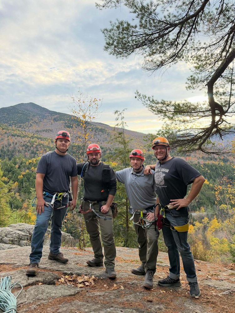 Four people wearing helmets and climbing gear stand on a rocky outcrop overlooking a hilly landscape with autumn foliage and a cloudy sky in the background.