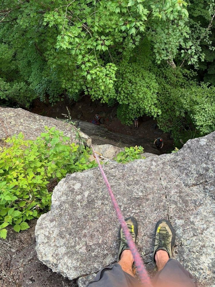 Feet wearing climbing shoes grip a rocky ledge as a rope stretches downward past trees into a forested area where climbers are visible below.