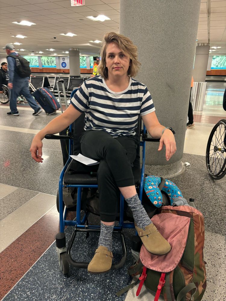 A white woman with blonde hair sits in a crappy airport wheelchair that she has to be pushed around in, despite being an athlete in town for a national tournament. The look on her face reflects her frustration.