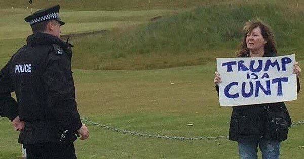 The late, great, Janey Godley holds a sign that reads “Trump is a Cunt” at the Orange asshole’s golf club in Scotland. A policeman looks on approvingly.