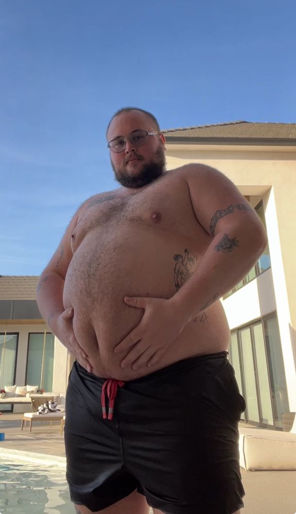 A 6 foot 350 lb man in a black swimsuit grasping his big belly in front of a pool.