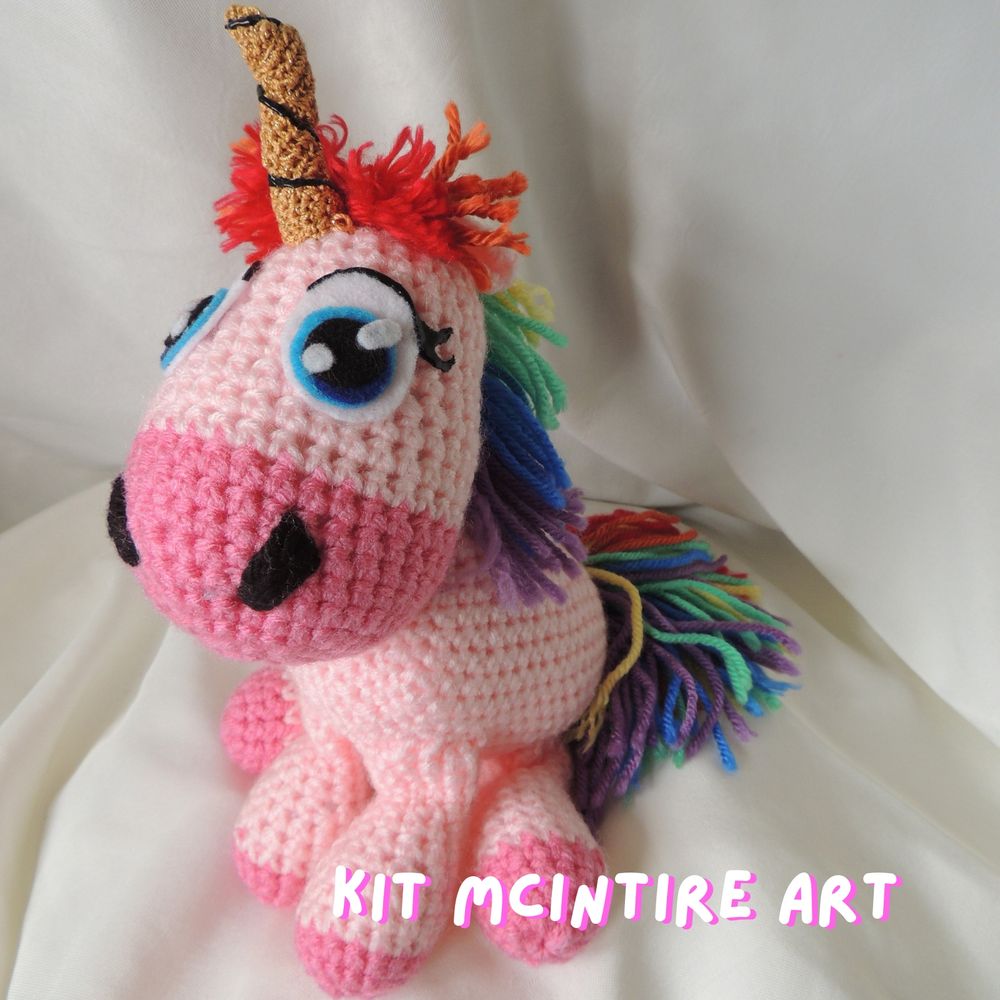 An original crochet unicorn plush. It has a rainbow mane and tail, a golden horn, and bright blue hand cut felt eyes.