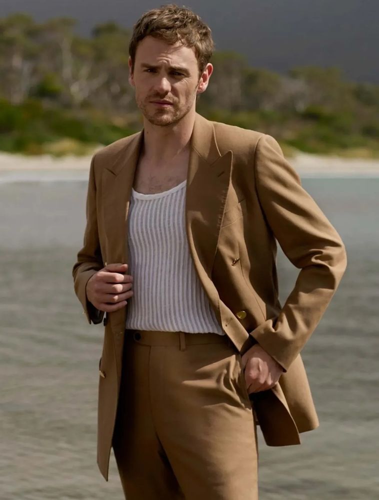 Charlie Vickers stands on a beach in a light brown suit. 