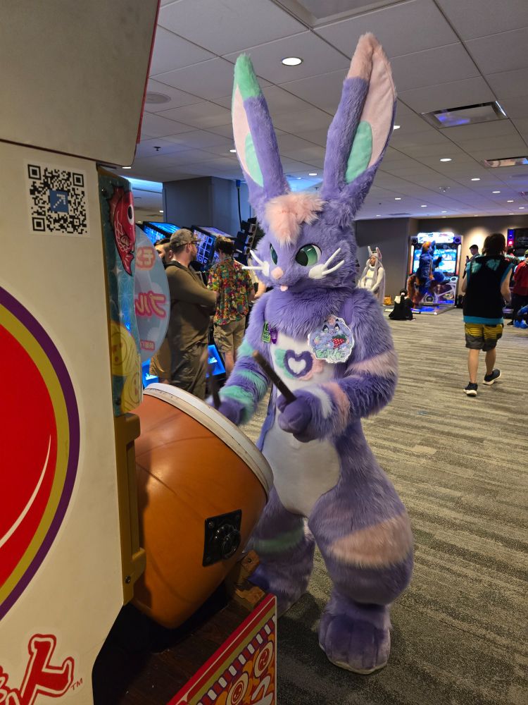 Purple bunny fursuiter playing a Taiko drum with drum sticks attached to a rhythm arcade game.