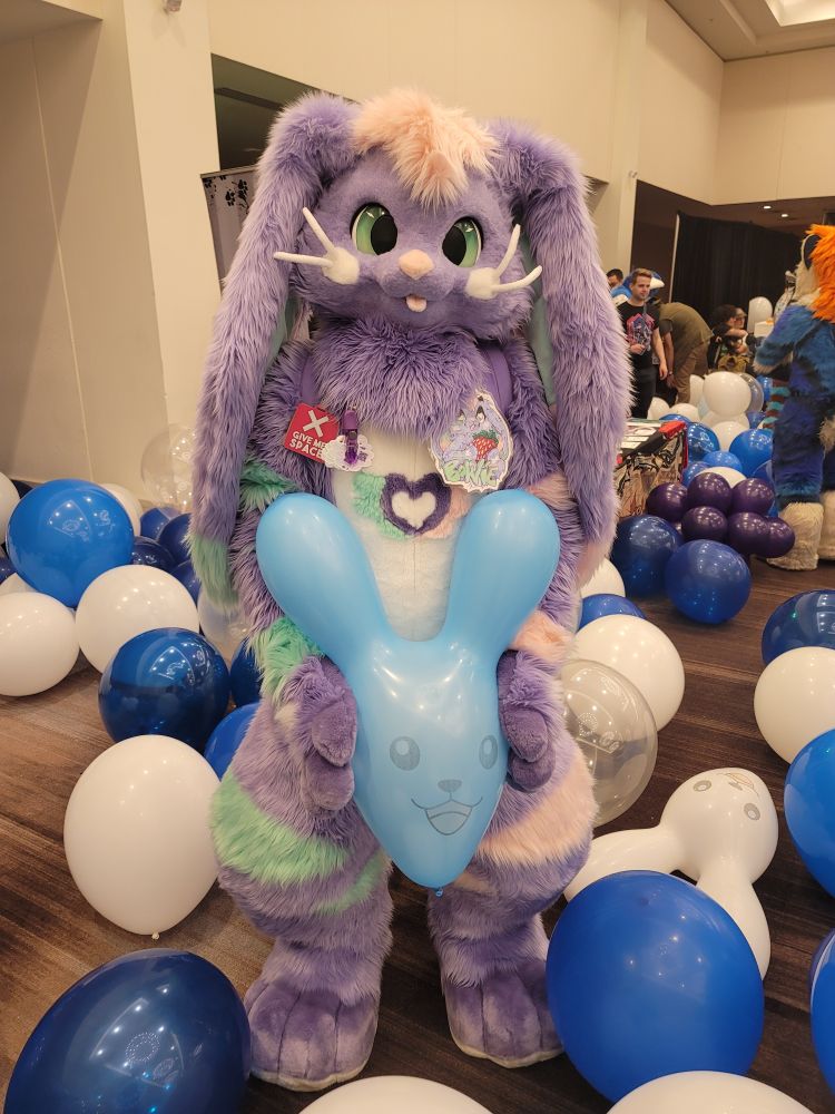 Purple bunny fursuiter holding a blue balloon in the shape of a bunny's face. They're surrounded by many other balloons and pool toys.