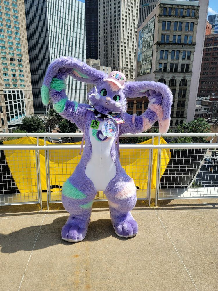 Purple bunny fursuiter holding their extra large lop ears up and out to look like wings. They're standing next to the edge on a balcony overlooking a city.