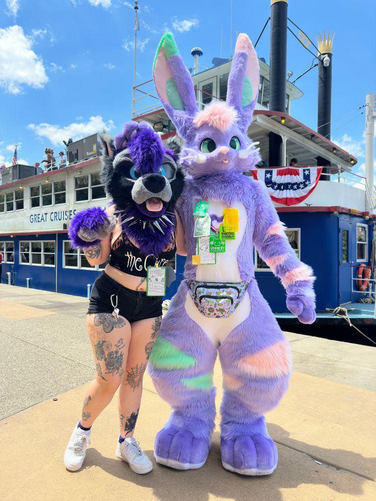 Purple bunny fursuiter posing next to another purple fursuiter on the waterfront at Anthrocon with the river cruise boat taking up the background