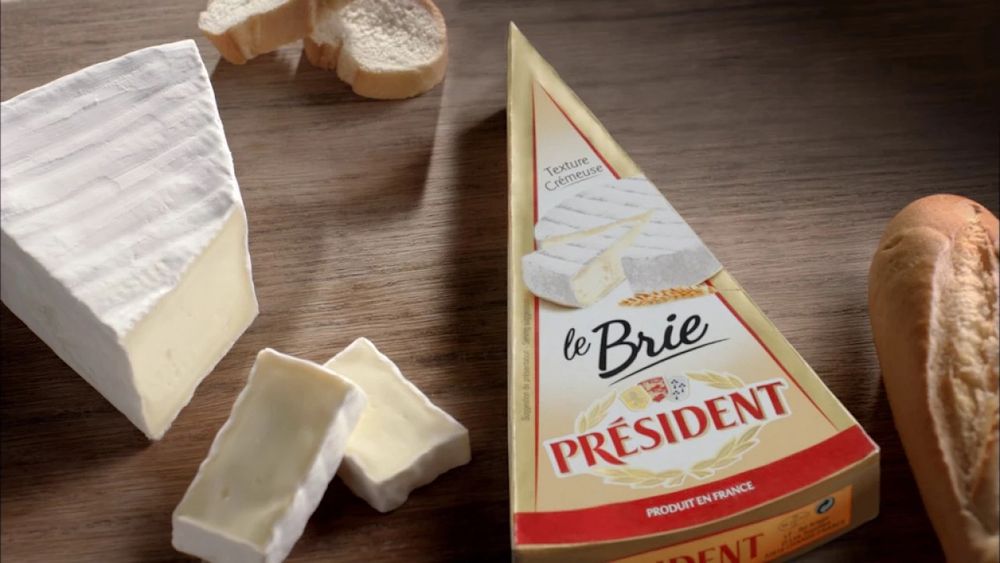 A wedge of President Brie, wrapped, plus an unwrapped and sliced wedge of the same. Placed on a wooden chopping board with a lovely French loaf.