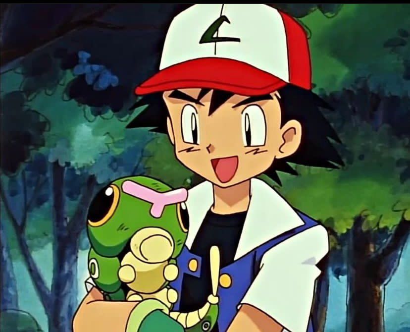 Ash Ketchum from the Pokémon anime, holding a green caterpillar called Caterpie in his arms. Ash has a big and relaxed smile on his face, and he looks at the Caterpie with adoration. The Caterpie is slightly curled up, but still looking at the viewer and Ash. The duo is standing in a forest clearing, with many trees in the background.