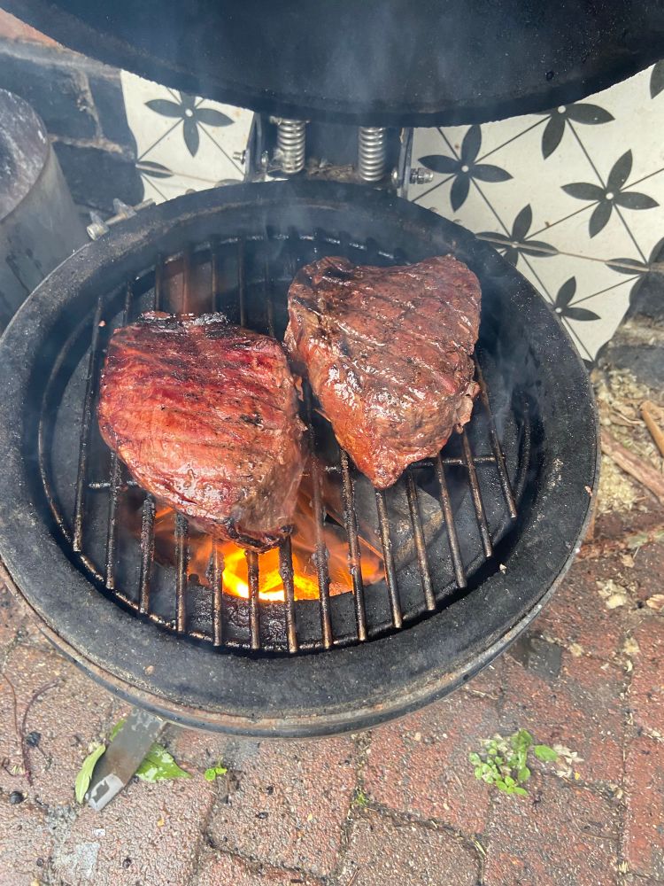 Two thick steaks cooking over a bbq grill with a flame underneath 