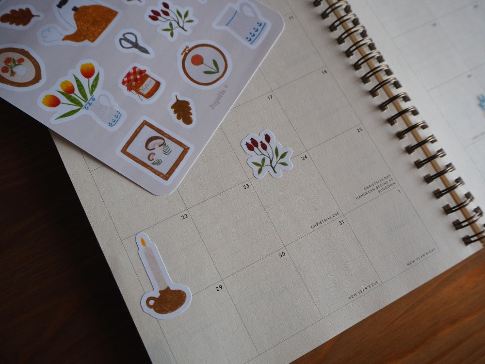Photo d'un agenda posé sur un bureau en bois. On a collé sur l'agenda deux stickers provenant d'une planche de stickers de ma boutique sur le thème cottagecore. Les deux stickers collés représentent une bougie haute dans un porte-bougie en métal cuivré, et une plante avec des bourgeons rouge foncé.