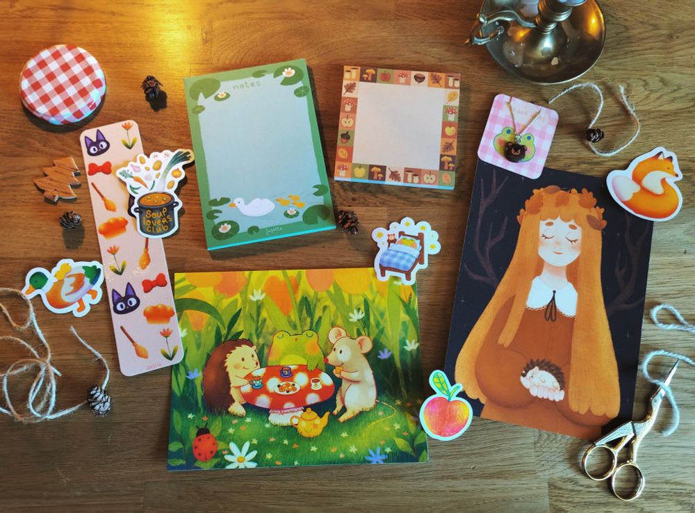 Photo de plusieurs articles de ma boutique mis en place sur un bureau en bois. On voit deux prints, un marque-page Jiji de Kiki la petite sorcière, deux bloc-notes, quelques stickers et des éléments de décoration (des fils de laine, des mini pommes de pin, des ciseaux cigogne dorés, une bougie en laiton et un couvercle de bocal à confiture.)