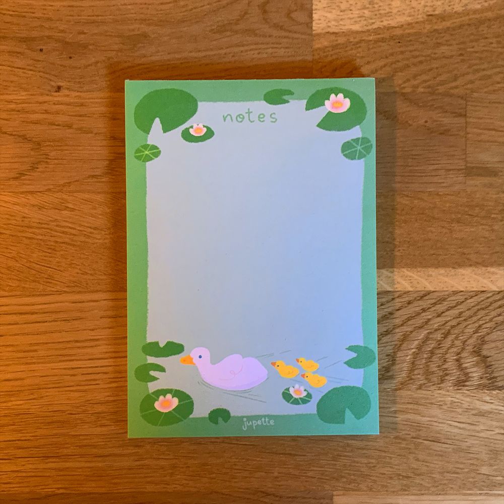 Un bloc-notes rectangulaire au format portrait est posé sur un bureau en bois. Le bloc-notes est vert clair et cyan pâle, et représente un canard blanc et ses petits canetons le suivant à l'arrière (les canetons sont jaunes) en bas du bloc. En haut, il est noté "notes" en vert.
Tout autour du contour du bloc, des nénuphars sont illustrés, certains avec une fleur de lotus.