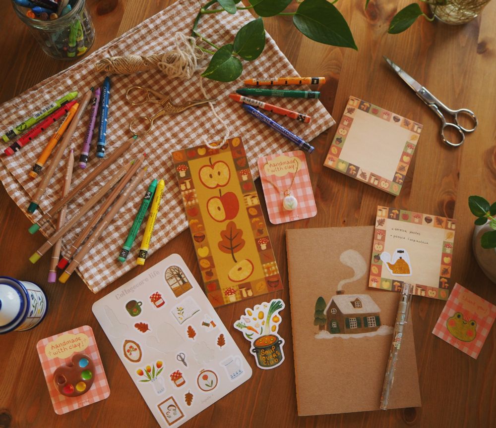 Photo de plusieurs articles de ma boutique mis en place sur une table en bois. On y voit un marque-page avec des pommes, un bloc-notes, une planche de stickers, un pin's en argile en forme de palette de peinture, un pendentif en argile blanche avec une fleur peinte sur le dessus et des éléments de décoration (des fils de laine, des ciseaux, crayons de couleur et pastels, un tissus vichy orangé brun, des plantes etc)