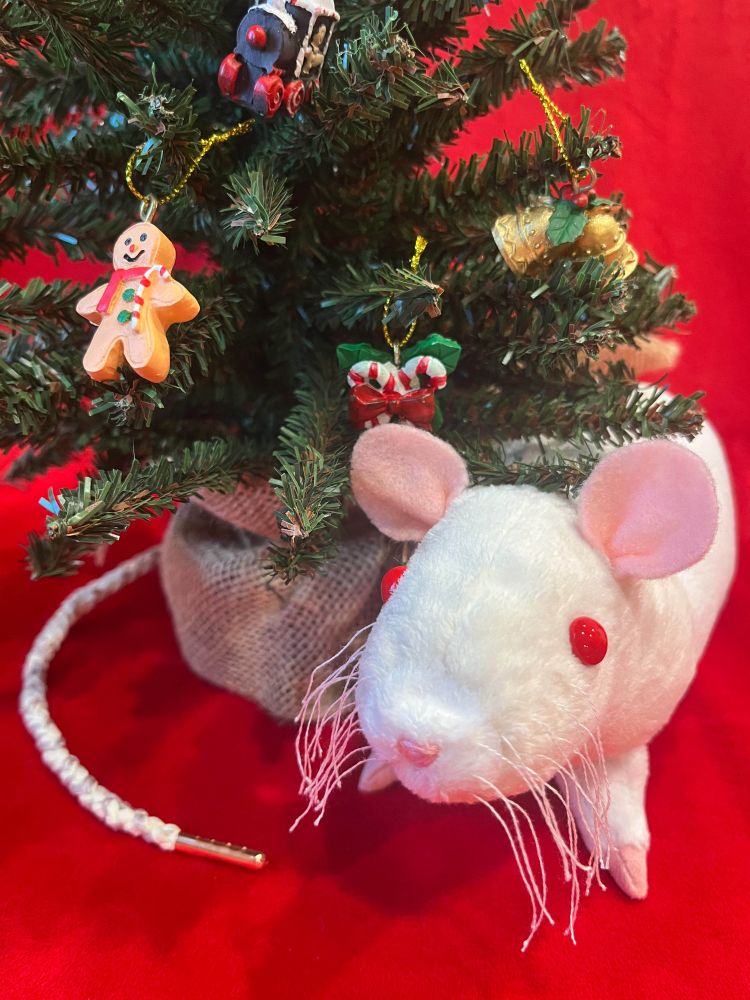 A cream-colored rat doll with a white face point is standing next to a small decorated Christmas tree. The doll has red eyes, and a braided cream-colored tail threaded with silver embroidery floss and tipped with a rose gold aglet.