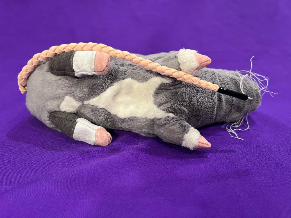 A gray rat doll with white markings on his tummy, wrists, and feet. His tail is a pale pink braid, tipped with a black aglet. He is lying on his side so his markings are facing the camera.