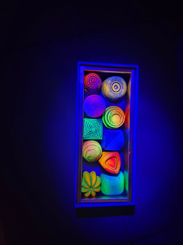 Assortment of chocolates under UV lights, making their colors extra vibrant. Each chocolate has a unique shape and color, from top to bottom and left to right: bright red and blue swirl, yellow and blue rings, purple and blue circle, green and yellow rings, blue and green squares that rotate, deep blue X, orange and yellow swirl, orange and red triangle, orange and green flower shape, and blue and green square.