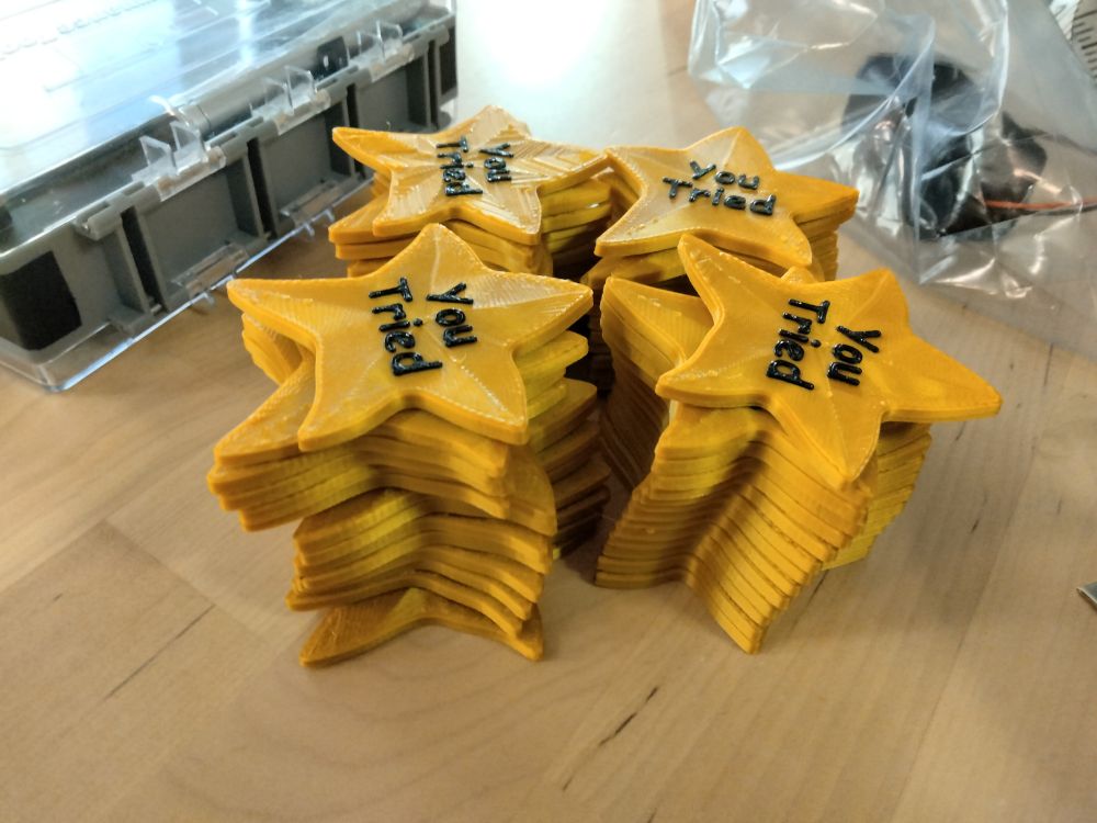 A stack of 3D printed lopsided gold stars with the text "You tried" printed on them in raised lettering.