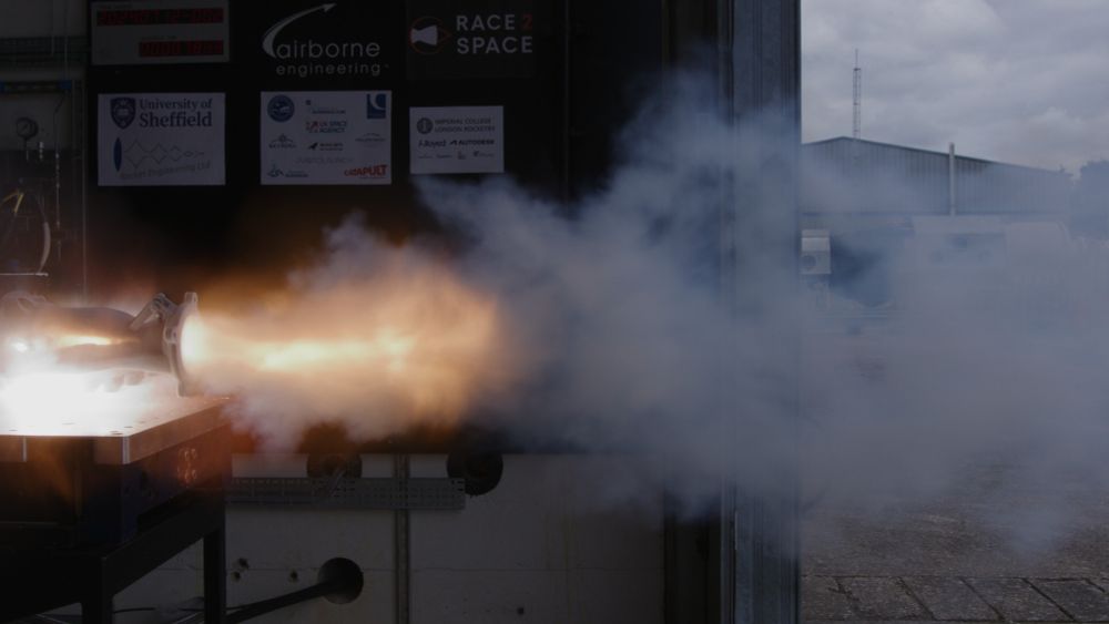 A photo of a small rocket engine being test-fired on a test bench. The engine has just started to explode and is in the middle of ripping itself apart, so a huge crack is tearing it open and revealing that the inside is full of light and fire.