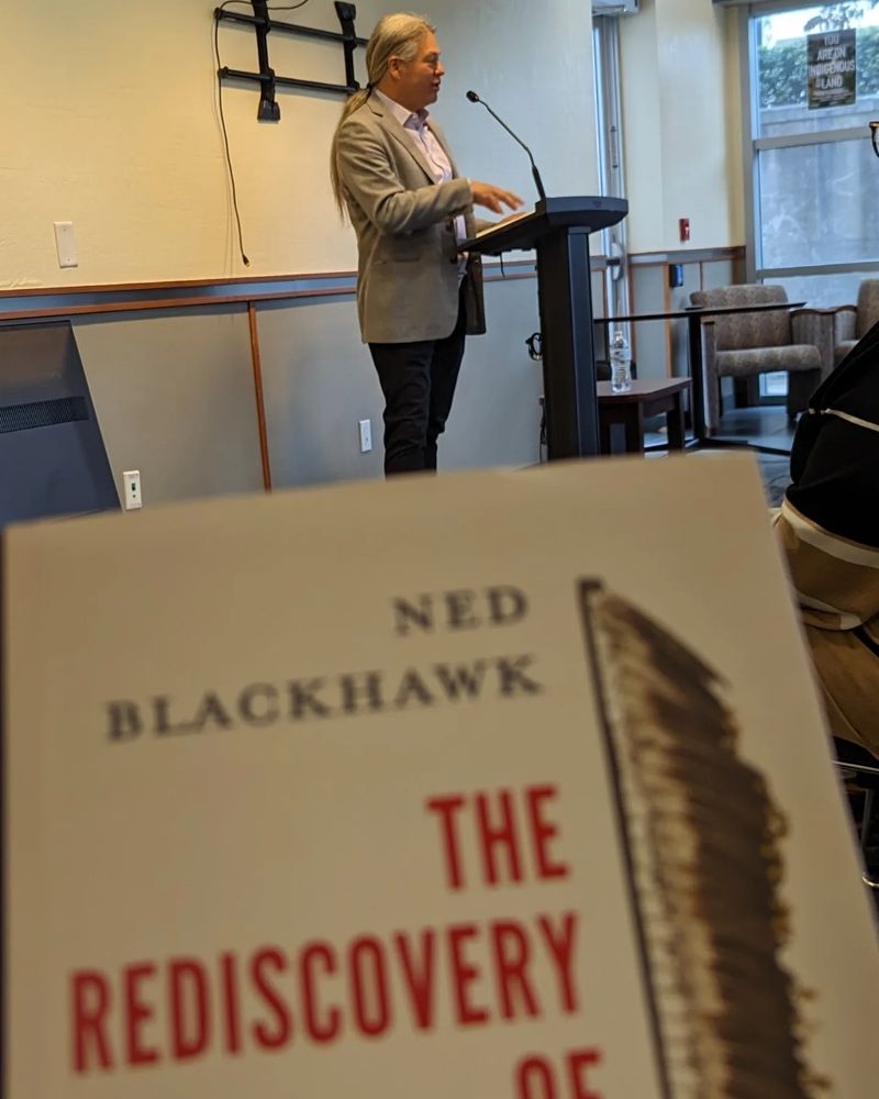 Book cover of The Rediscovery of America, Native People and the Unmaking of US History with Ned Blackhawk, a Native man with long white hair, speaking at a podium behind the book.