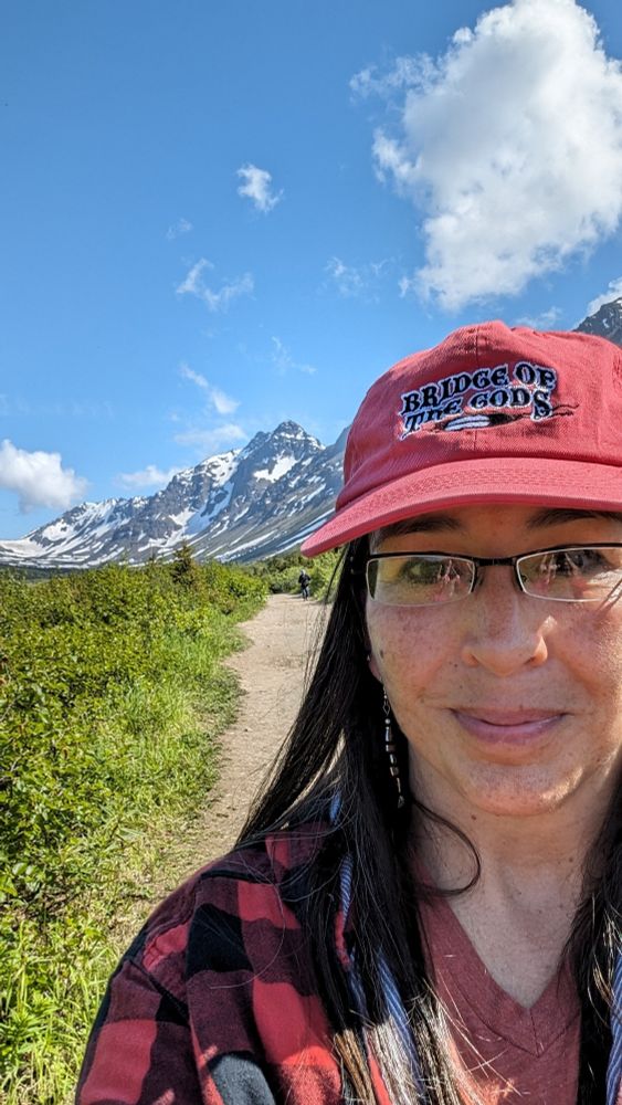 A smiling woman wearing a red baseball hat that says Bridge of the Gods stands on a dirt trail. A line of mountain peaks rises above her right shoulder