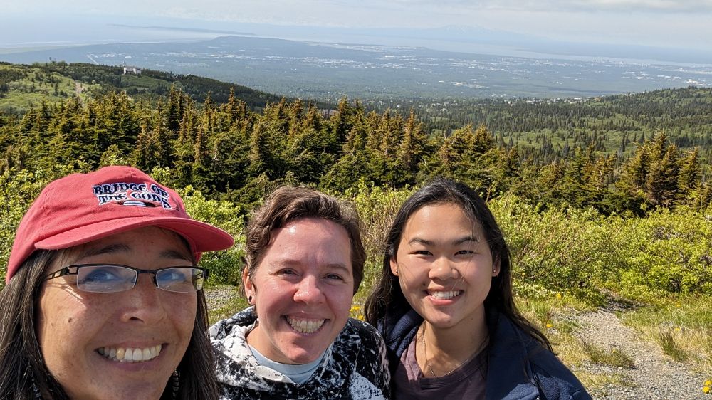 Three people smiling with a green mountain side behind them.a city is just barely visible through the mist below