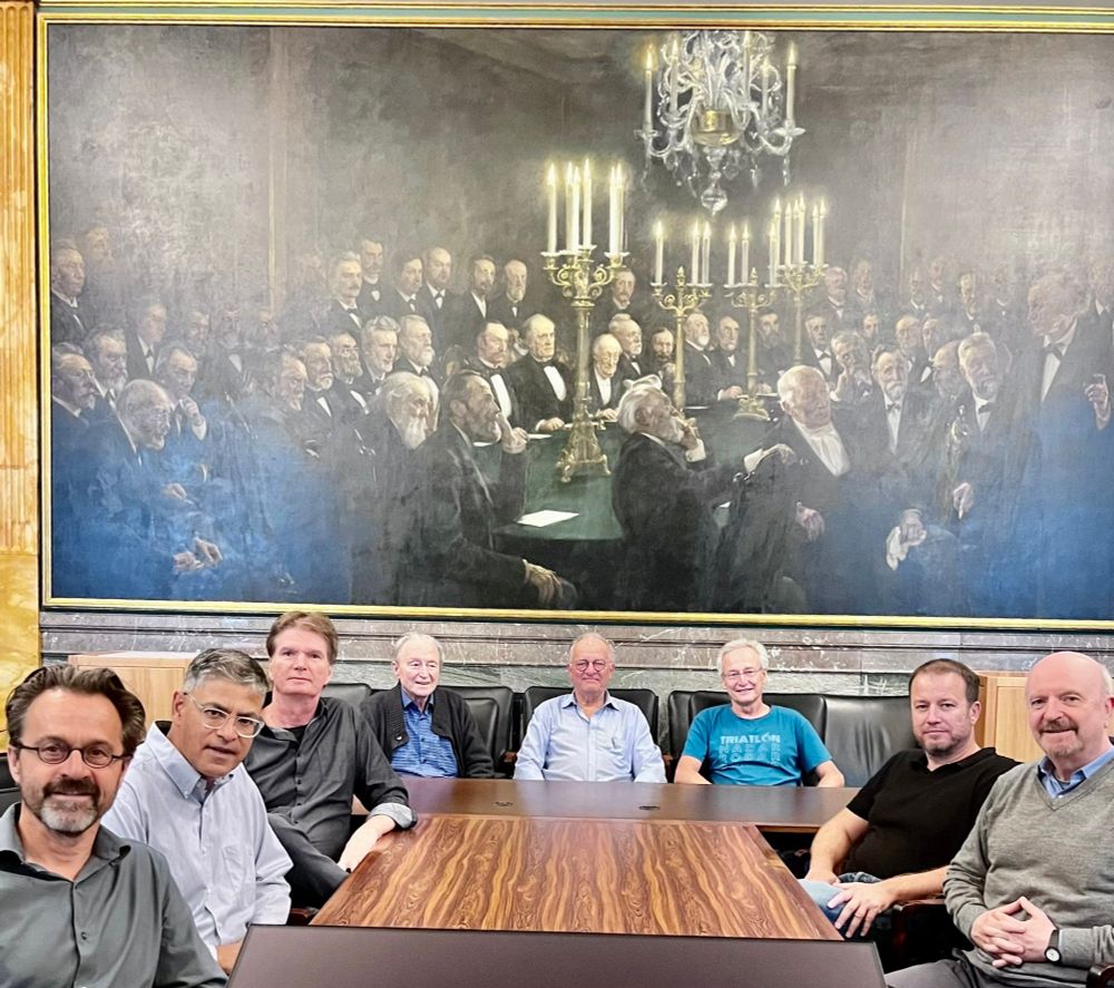Photo of a table of mostly white men sitting in front of a very large painting of white, mostly elderly, men