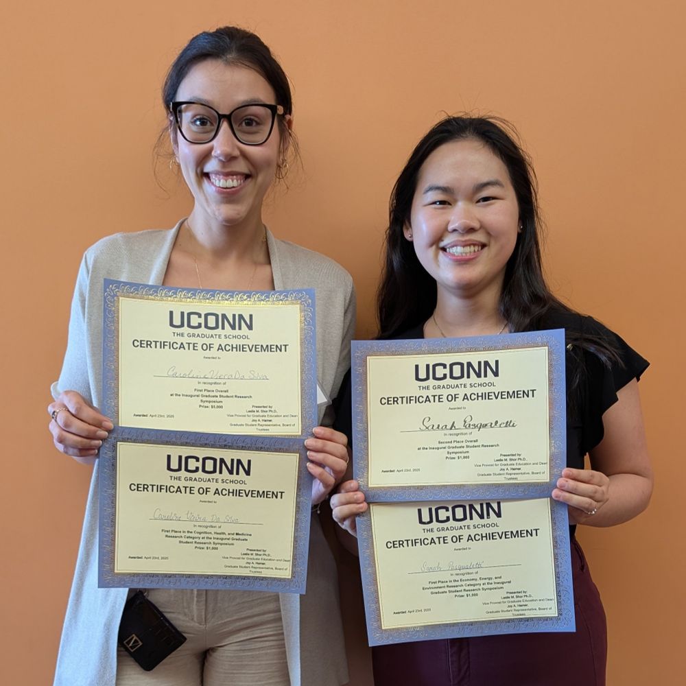 2 women stand smiling and holding certificates of achievement for winning first place in their categories and first and second place overall in the poster competition. One is wearing glasses and tan sweater and pants, and the other wears a black shirt sleeved shirt and maroon pants.