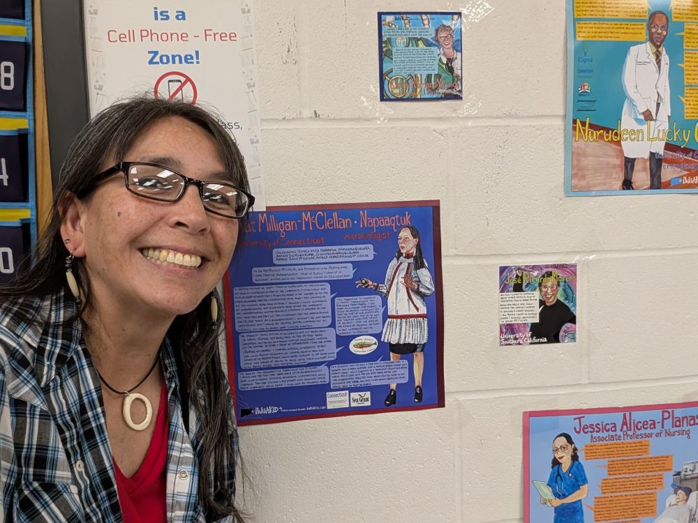 Smiling woman wearing a blue checkered shirt and ivory jewelry stands next to a poster with a cartoon of herself and a description of her work and some quotes. 4 other posters in similar design but of other scientists and other professionals are also on the wall