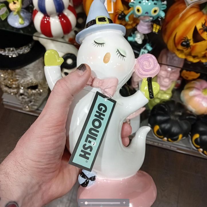 a cutesy ceramic ghost in a blue witch's hat holding a pink lollipop - it has a tag with the word "GHOULISH" on it