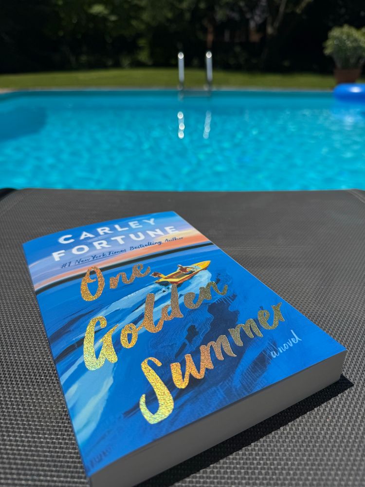 Cover of the book ‘One Golden Summer’ by Carley Fortune beside a pool 