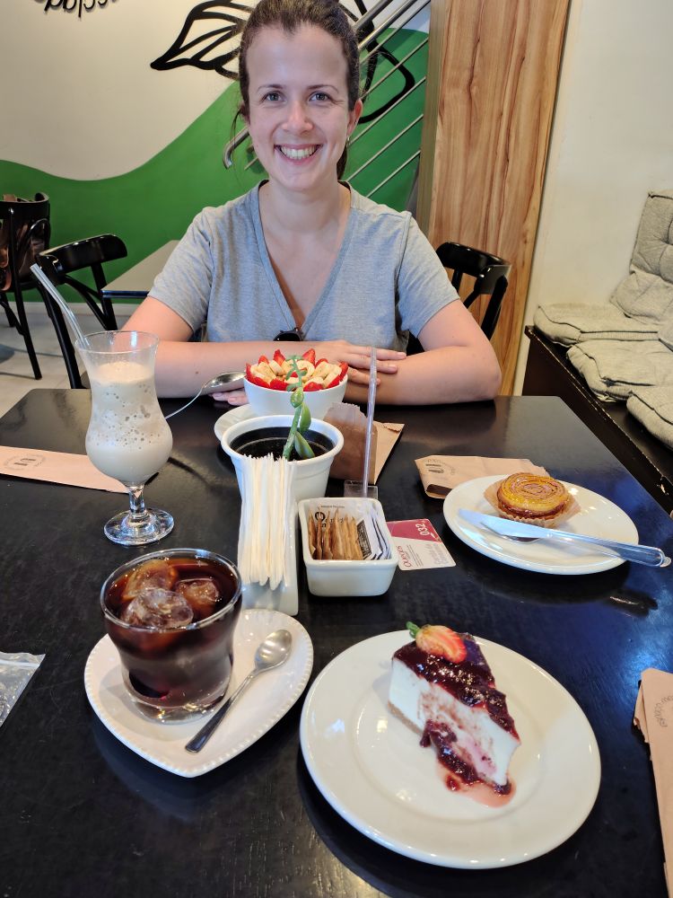 A foto mostra um momento agradável em uma cafeteria. Na imagem, minha esposa está sorrindo à mesa, vestindo uma camiseta cinza e com um semblante alegre. À frente dela há uma tigela com açaí, decorada com morangos e bananas. Sobre a mesa, há também várias delícias: uma fatia de cheesecake com calda de frutas vermelhas e um morango por cima, um copo de café gelado com gelo, um milkshake claro, e uma pequena torta sobre um prato ao fundo. 
