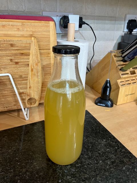 A glass bottle almost full of a yellow-green liquid standing on a kitchen worktop.