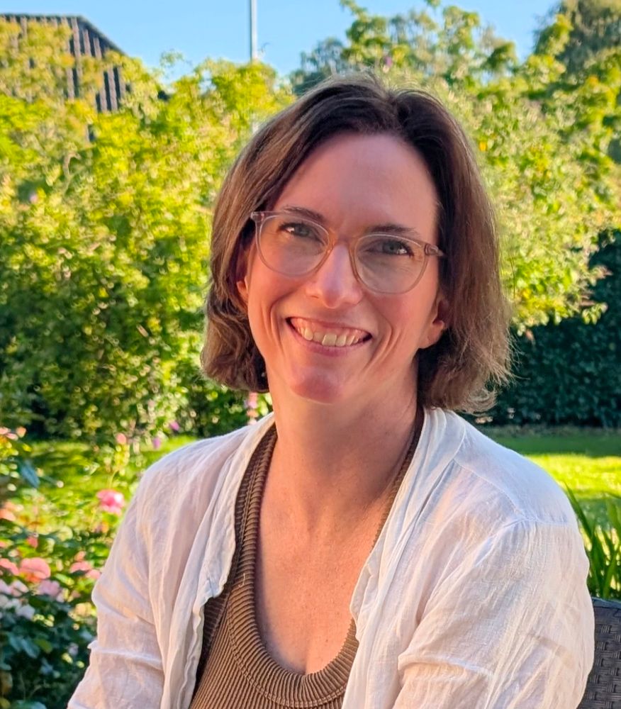 Sumar-Halla brosandi í hvítri opinni skyrtu og brúnum bol, með brúnt axlasítt hár og gleraugu 