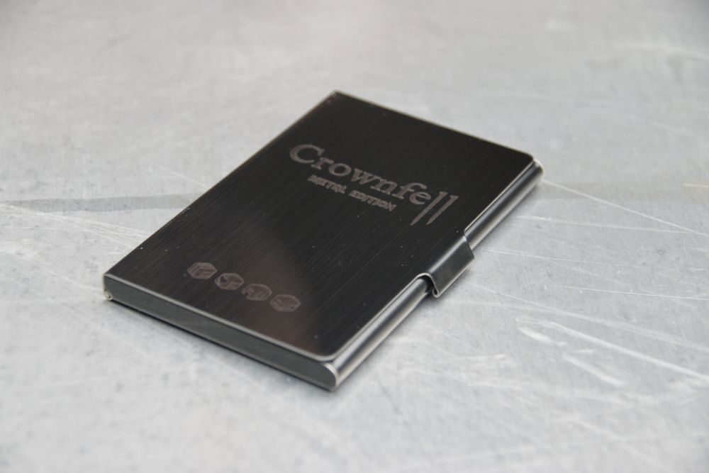 The metal Crownfell wallet, sitting on a sheet of aluminum. It's a black steel wallet with the logo laser etched on the cover.