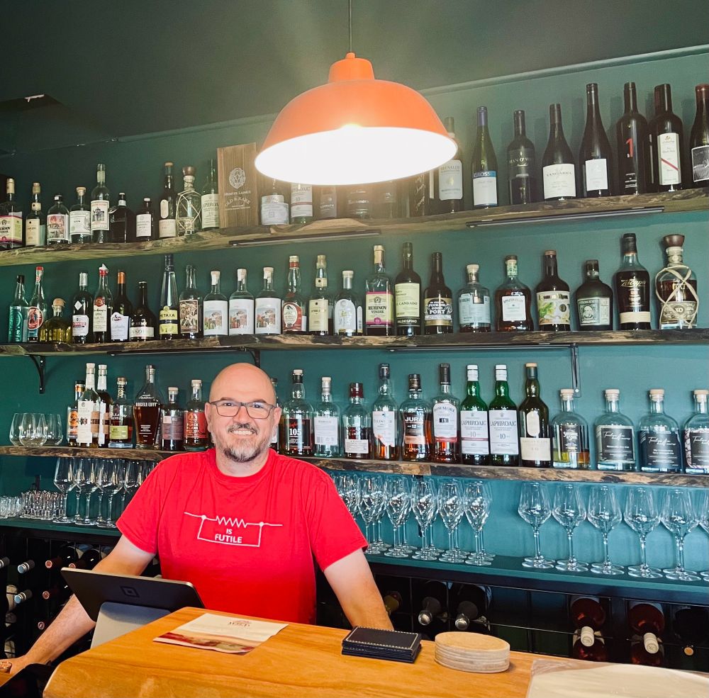 A man in a red shirt stands smiling behind a bar with wine and whisky bottles and glasses on the wall behind him