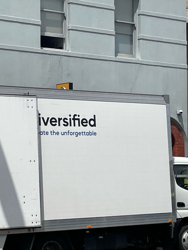A white van on the side of the road. The back doors are open, partly obscuring the sign on the side, which reads: 
-iversified 
-ate the unforgettable 