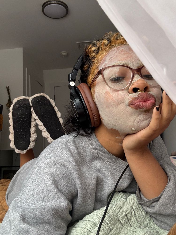 Creator x lays in bed looking comfy with headphones and cozy slippers with a face mask on