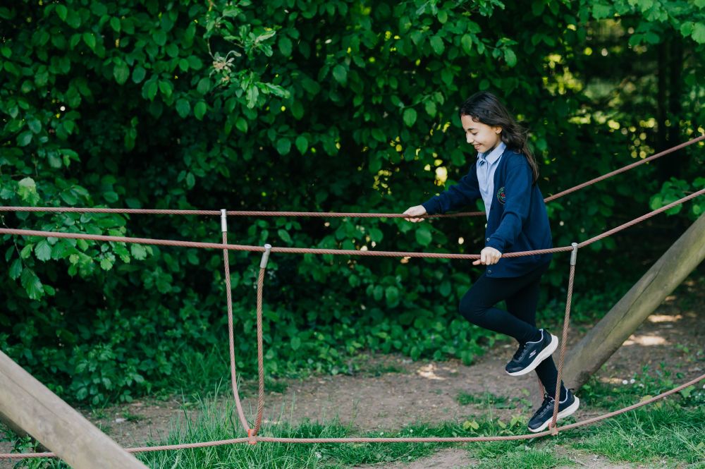 A school child walks along a rope bridge. They are wearing a light blue polo shirt, a dark blue cardigan, black leggings and black trainers. 