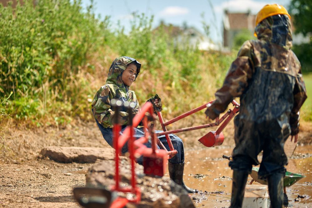 A photo of two children playing outdoors in a mud pit on a school field. They are wearing head to toe waterproofs and are using a play digger. 