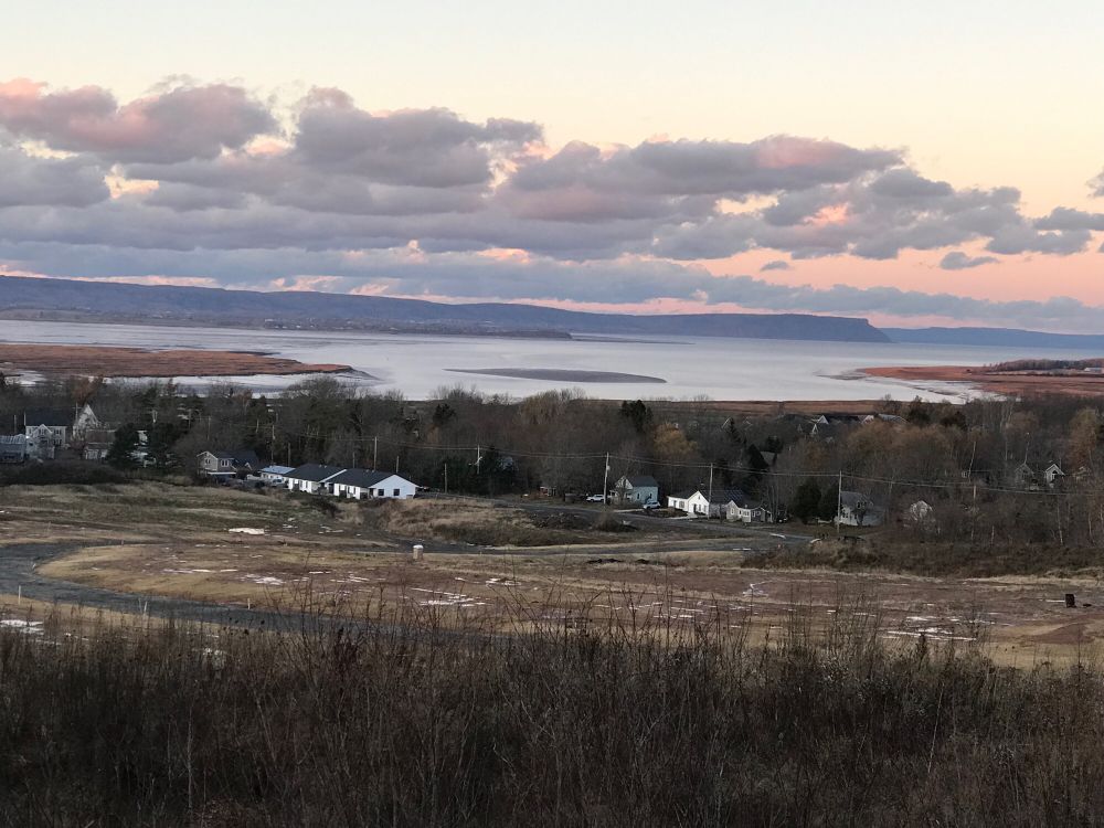 A photo of the Minas Basin, with the North Mountain and Blomidon in the distance. A field and a few houses are in the foreground. The sky is partly cloudy and pinkish from the late afternoon sun.