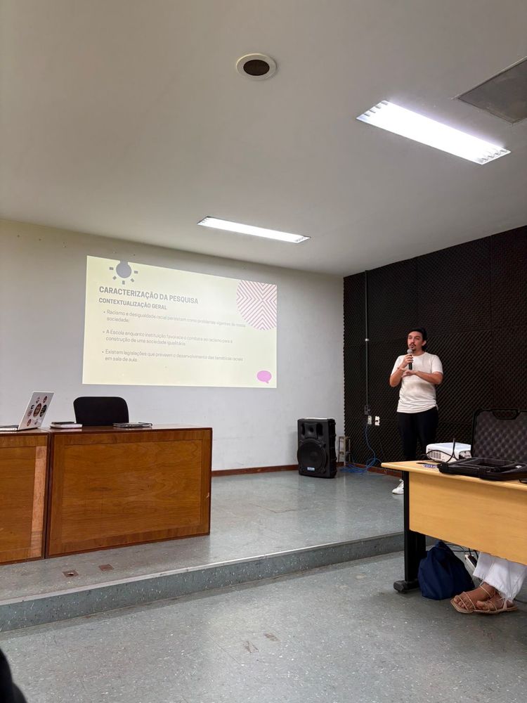 eu, homem branco, cabelos na altura do pescoço, pretos. cavanhaque e bigode preto. foto de corpo inteiro. uso uma camisa clara, uma calça social e um tênis da adidas branco com três linhas pretas. seguro um microfone e gesticulo enquanto falo.