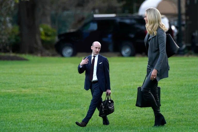 A photo of a bald man and a blonde woman both in business attire walking across a lawn, the man is considerably shorter than the woman