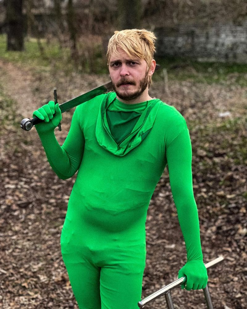 Holden relaxing in the woods resting a sword on his shoulder, with a sad look on his face. Wearing a skin tight green body suit with the hood down.