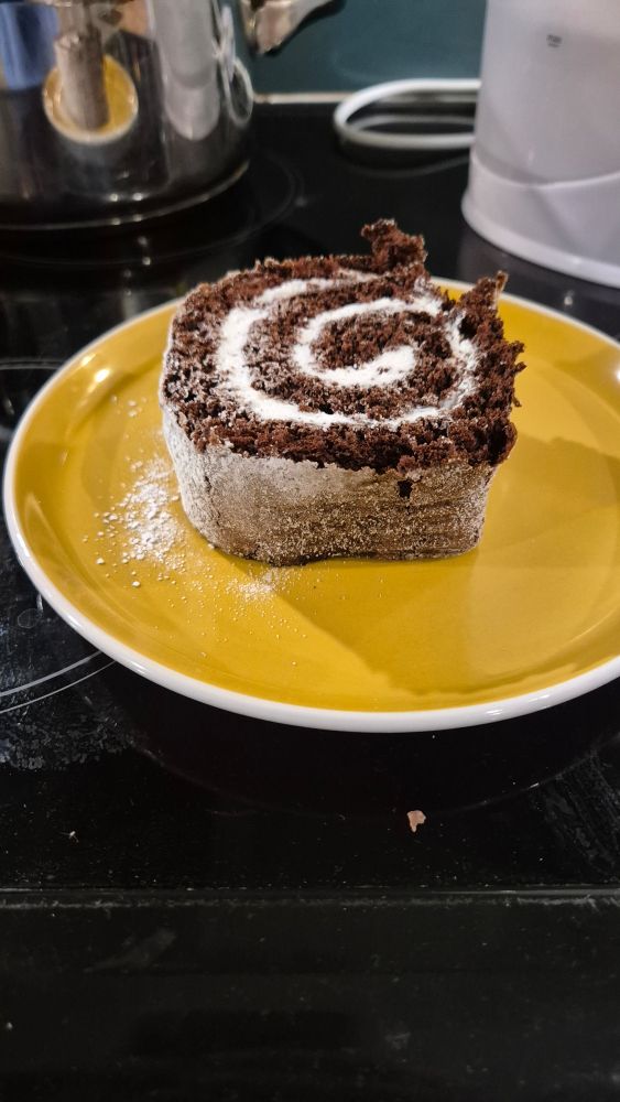 The black top of a hob, with a yellow plate central bearing a large slice of chocolate swiss roll. 