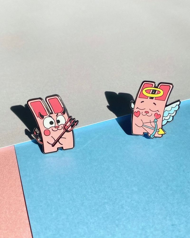 The Sims Freezer-bunny themed enamel pins, two make up a Devil/Angel duo and they are placed against blue and pink paper in this image.