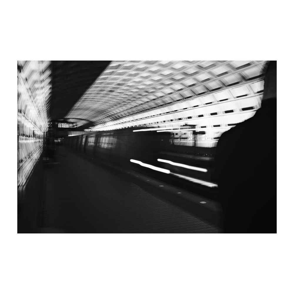 an abstract black and white image of blurred, trailing lights from public transportation cut in thirds by shadow and flanked by bright geometric grid patterns. the silhouette of a traveler sits to the right of the frame