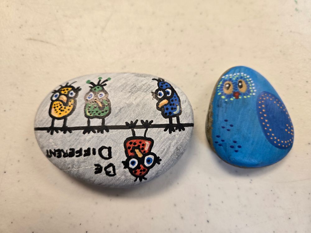 Two small flat stones.

One is painted white with three birds (yellow, green, and blue) perched on a line, while a fourth red bird hangs upside down. Text beside the red bird, also upside down, reads "Be different."

The other is painted a blue bird, either yellow eyes surrounded by thin lines and dots, and a dark blue wing with yellow dots along the edge.