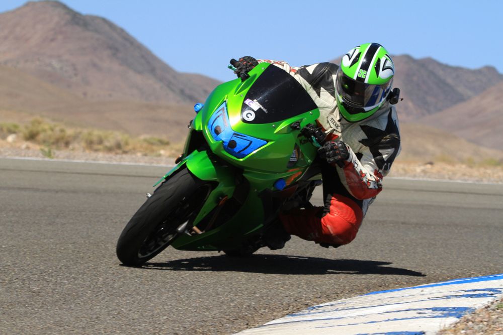 A green Kawasaki Ninja 250 on a race track out in the dry brown desert. The rider is in white and red leathers with black trim, a full face neon green helmet with white and black accents. She is leaning into a curve, knee out (though definitely not touching) facing straight toward the cameraman who is kneeling low on the inside of the curve. The headlight lens is taped over and a pair of angry eyes are sharpied onto the blue tape, making the bike glare ahead at the track. There's two C stickers on the black windscreen, showing this is part of the slow group. 